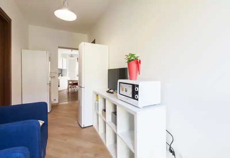 Espace de vie compact avec un fauteuil bleu, vue ouverte sur la cuisine et étagère avec micro-ondes et télévision — agencement lumineux et fonctionnel, parfait pour de courts séjours.