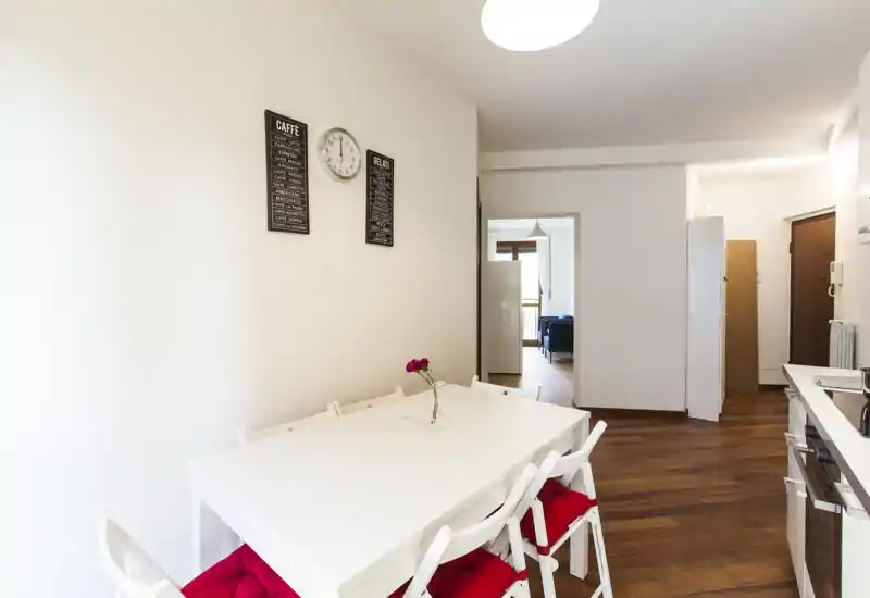 Espace repas lumineux et moderne avec une table blanche et des chaises pliantes avec coussins rouges, un vase avec une fleur et des décorations murales; vue dégagée vers le couloir et le séjour adjacent.