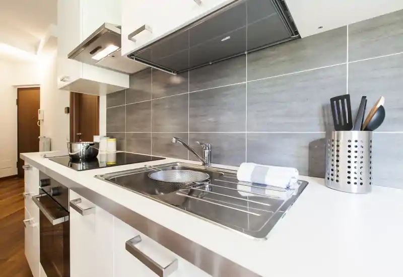 Cuisine compacte et moderne avec évier en inox, plaque à induction et ustensiles rangés; surfaces lumineuses et crédence grise contemporaine.