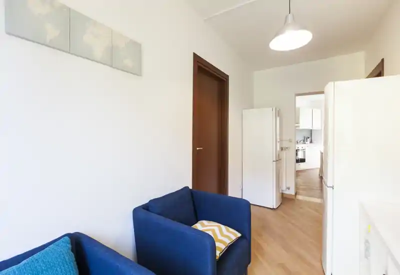 Petit salon avec fauteuils bleus en tissu, sol en bois et murs neutres clairs ; une porte donne sur la cuisine, espace commun pratique et lumineux.