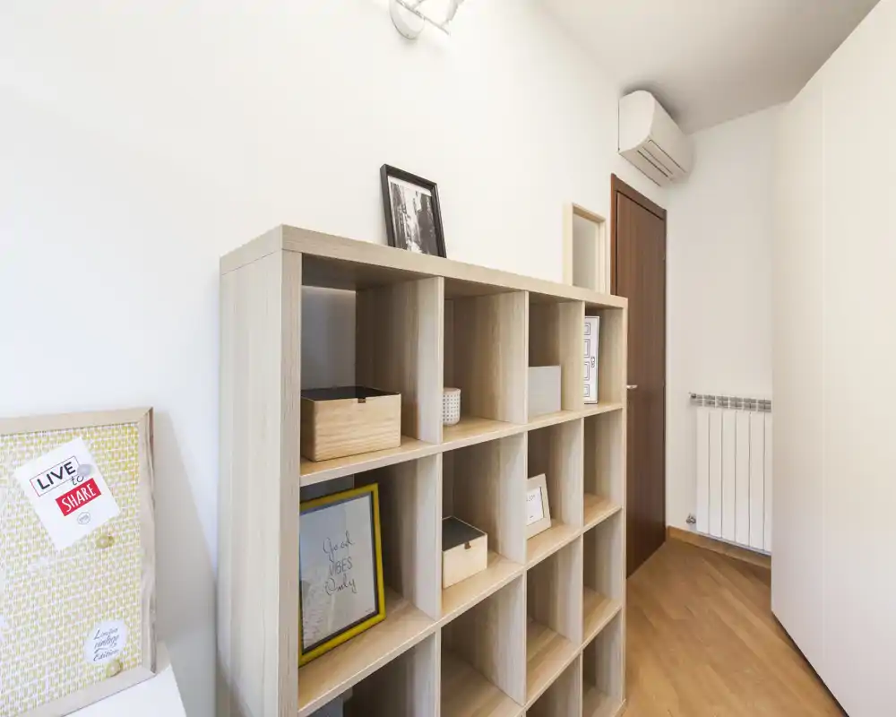 Rincón de estar luminoso y minimalista con estantería de cubos en madera, marcos decorativos y suelo de madera cálida: un recibidor/espacio de salón ordenado y moderno.