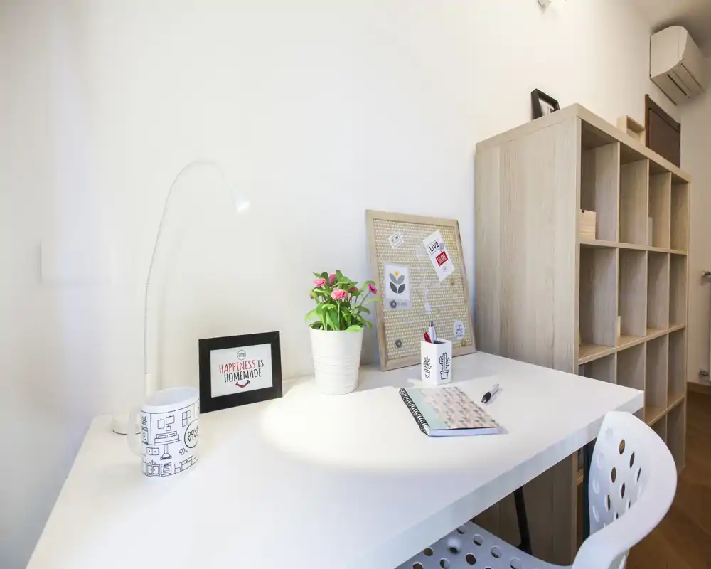 Espacio de trabajo en casa luminoso y ordenado con escritorio blanco, lámpara, planta y un tablero de corcho decorativo — una zona tranquila para trabajar o estudiar.