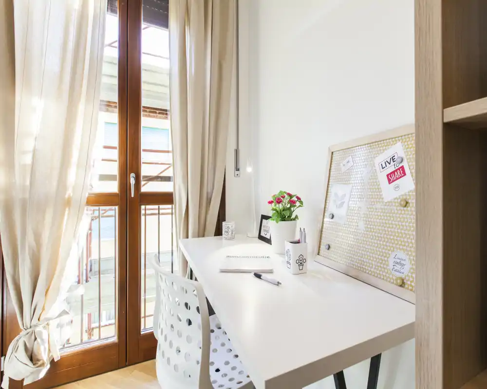 Espacio de trabajo luminoso junto a la puerta del balcón con un escritorio blanco ordenado, silla perforada, tablón de notas y una pequeña planta — ideal para teletrabajo con mucha luz natural.