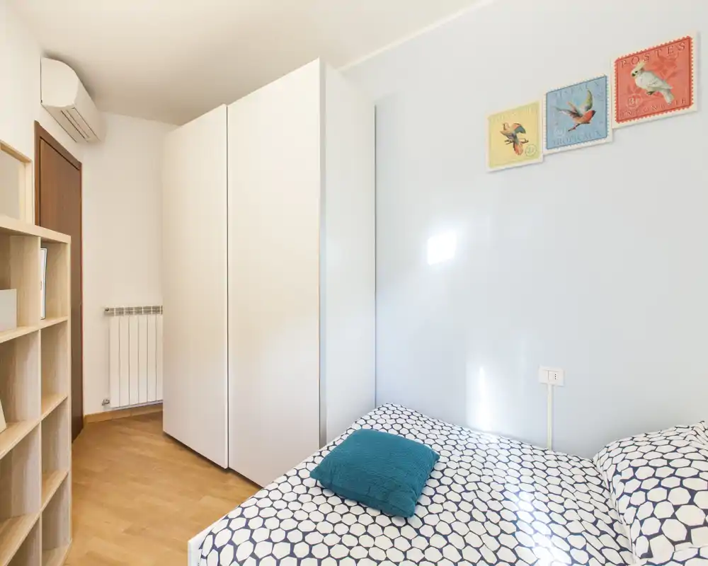 Dormitorio luminoso y bien presentado con cama doble estampada, cojín turquesa y un gran armario blanco; suelo de madera y pared en tonos pastel crean un ambiente tranquilo.