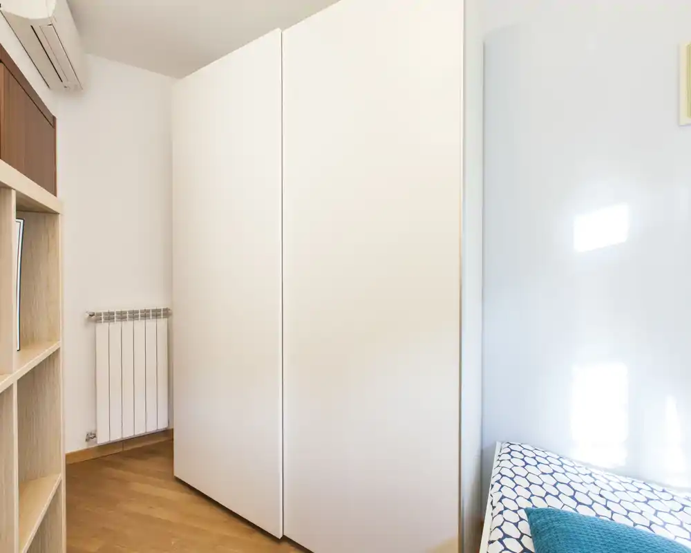 Rincón de dormitorio con un gran armario blanco y un lateral de la cama individual con colcha estampada; luz natural intensa y suelo de madera limpio.