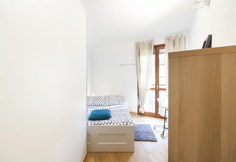 Dormitorio compacto y luminoso con cama doble con ropa de cama estampada, cojín turquesa, pequeña alfombra y puerta-balconera que aporta mucha luz natural.