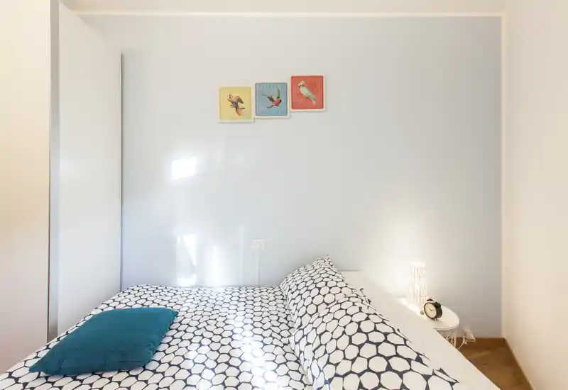 Dormitorio moderno y luminoso con cama doble vestida con ropa de cama estampada, cojín turquesa y luz de mesita cálida — ideal para una estancia acogedora.