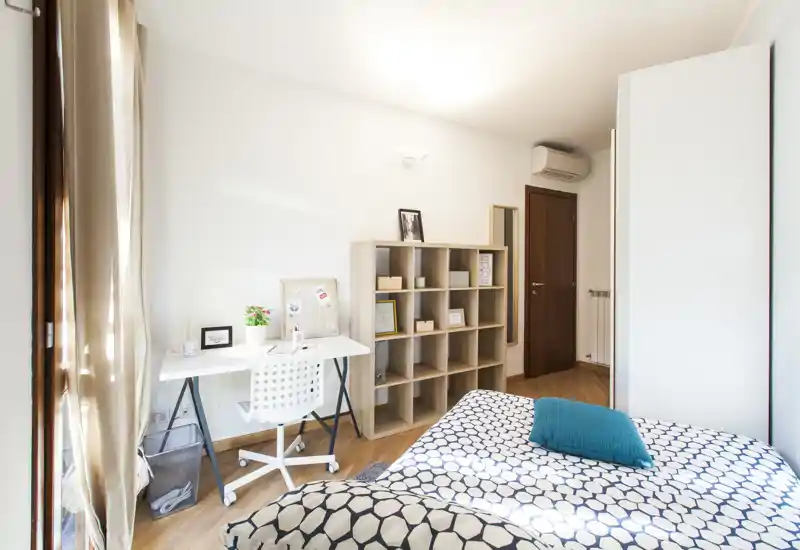 Dormitorio moderno y luminoso con colcha de estampado, cojín turquesa, escritorio junto a la ventana y estantería abierta: un espacio acogedor y bien iluminado, ideal para estudiante o persona sola.