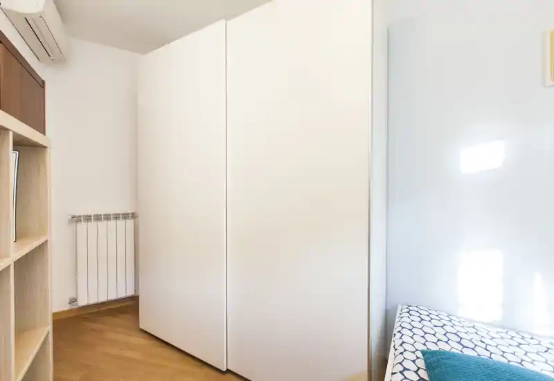 Rincón de dormitorio con un gran armario blanco y un lateral de la cama individual con colcha estampada; luz natural intensa y suelo de madera limpio.