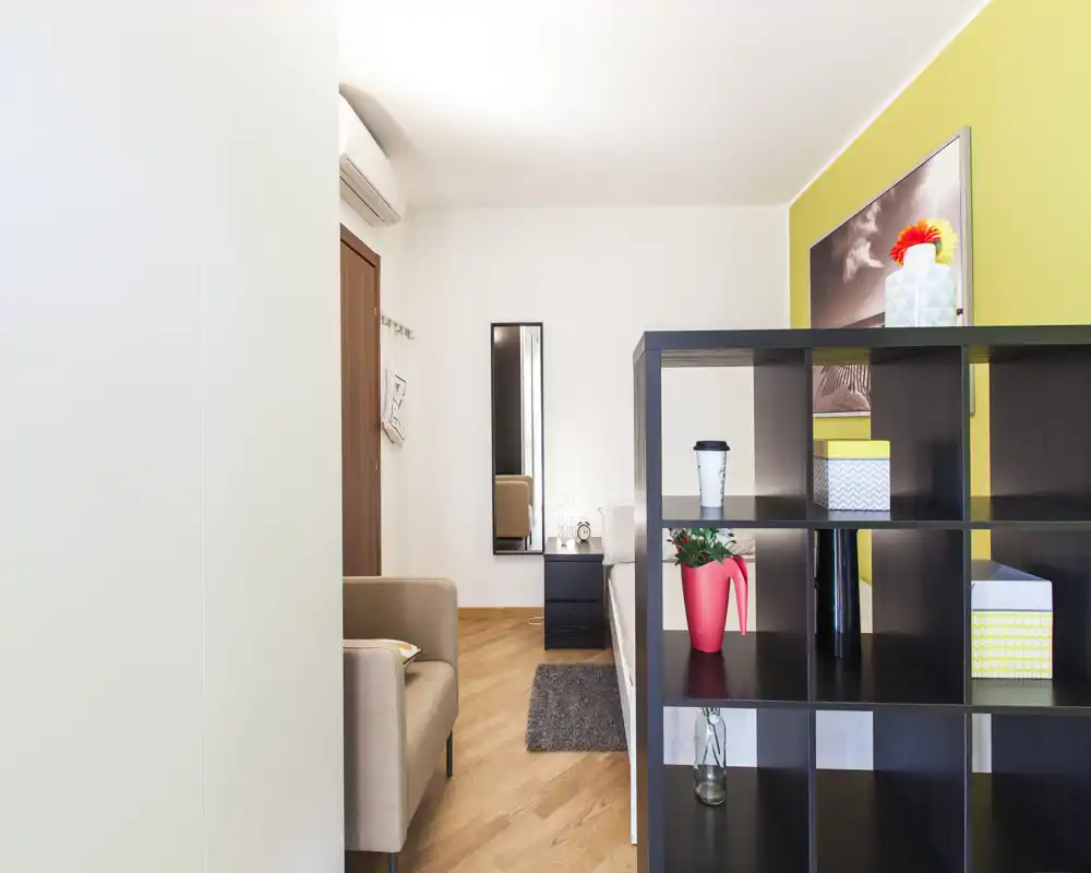 Zona giorno luminosa e ben arredata con una poltrona confortevole, scaffalatura decorativa usata come divisorio, un piccolo tappeto e dettagli curati—perfetta per un monolocale accogliente.