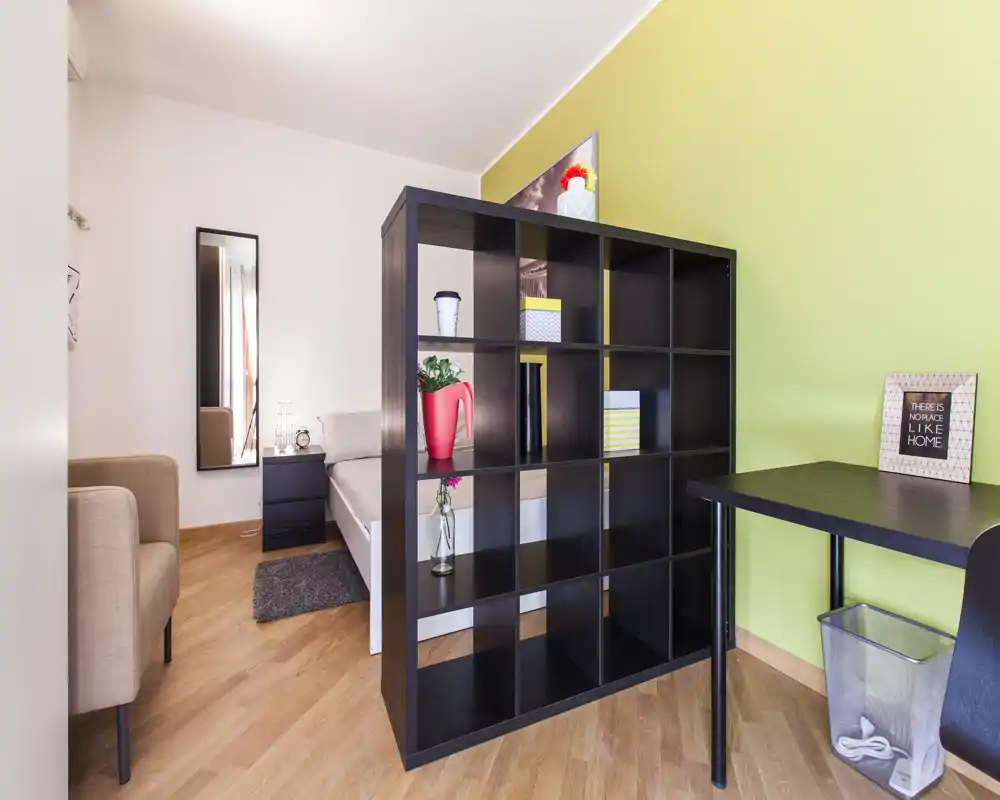 Camera da letto luminosa e ben arredata con un divisorio a scaffali moderno, letto matrimoniale visibile, comodino e tessili accoglienti — perfetta per un appartamento da uno o due persone.