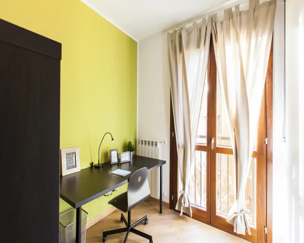 Ambiente luminoso e multifunzionale con scrivania nera e sedia ergonomica accanto a grandi finestre a tutta altezza con tende leggere — uno spazio lavoro accogliente e ben illuminato.