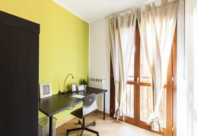 Pièce lumineuse et multifonctionnelle avec bureau noir et chaise ergonomique près de grandes fenêtres du sol au plafond avec voilages — un espace de travail chaleureux et bien éclairé.
