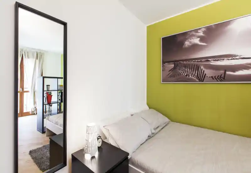 Compact, well-lit bedroom with a double bed dressed in neutral linens, a bedside table with lamp, and decorative wall art; a tall mirror and doorway add depth and natural light.