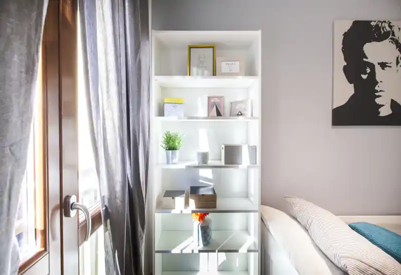 Rincón de dormitorio luminoso y moderno con una estantería blanca, ropa de cama suave y luz natural de una gran ventana. La imagen destaca el almacenamiento y el estilo ordenado.