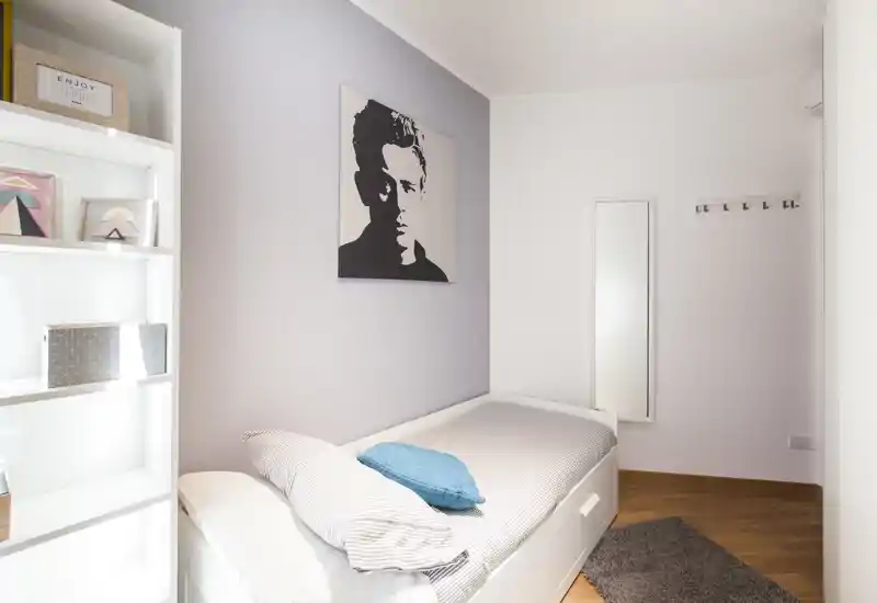 Dormitorio individual compacto y luminoso con cama bien tendida, cojín decorativo y cuadro en la pared. Líneas limpias y suelo de madera cálida que crean un ambiente moderno y acogedor, perfecto para estancias cortas.