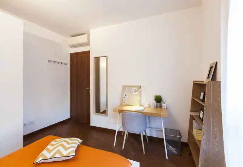 Dormitorio soleado y ordenado con cama en tonos naranjas, un pequeño escritorio de madera con lámpara y panel de notas, y una estantería — ideal para una estancia confortable o trabajo remoto.
