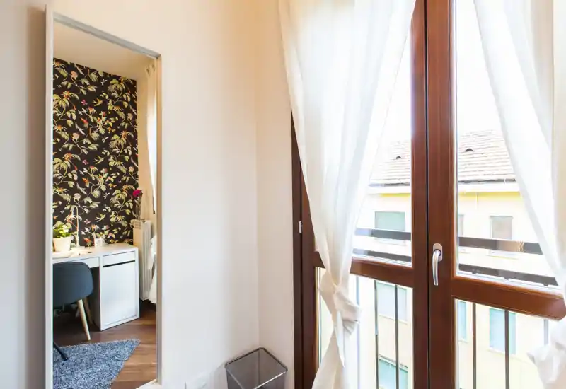 Rincón luminoso de una habitación con gran ventana y cortinas traslúcidas; la puerta muestra una zona decorada con escritorio pero no se ve la cama directamente.