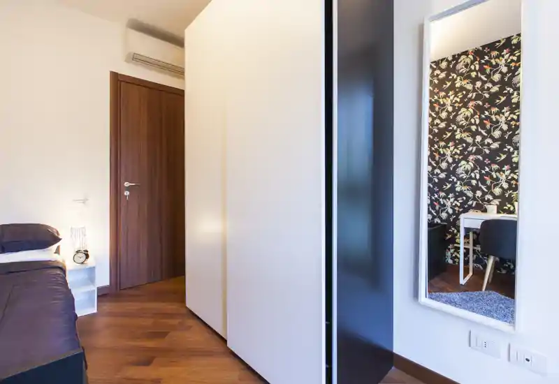 Rincón de dormitorio moderno y luminoso con cama individual visible, mesita de noche y suelo de madera cálido; un espejo de cuerpo entero refleja una pared decorada con escritorio.