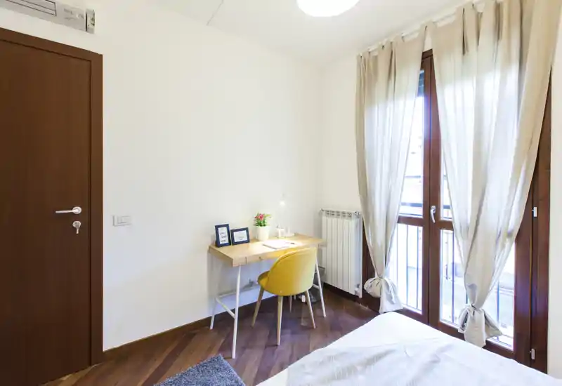 Rincón de dormitorio luminoso y acogedor con vista parcial de la cama, un escritorio de madera y una silla amarilla junto a grandes ventanas con cortinas claras.