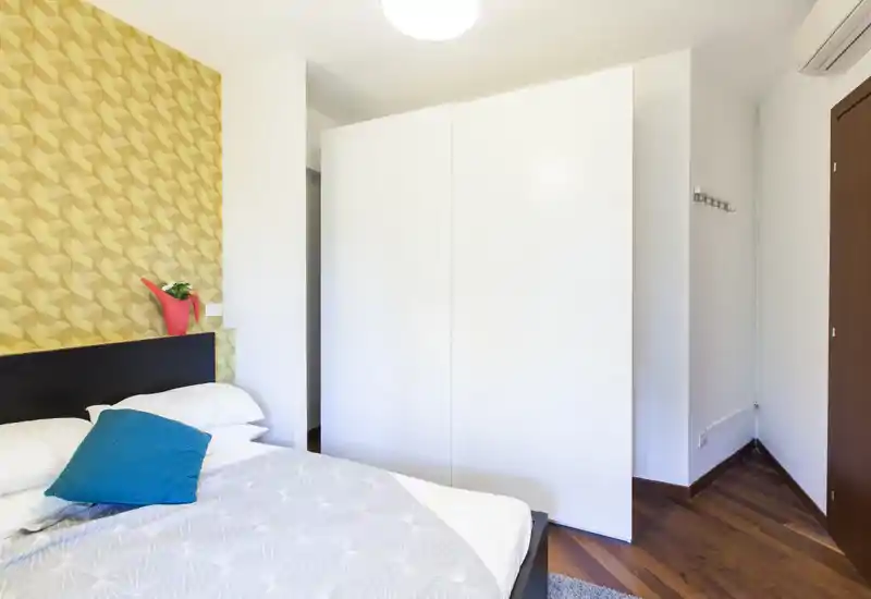 Dormitorio moderno y luminoso con pared decorativa, cama bien hecha y armario blanco limpio: ideal para anuncios de alquiler que destaquen comodidad y estilo.