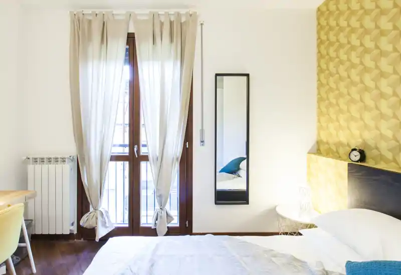 Dormitorio luminoso y moderno con la cama bien hecha, pared amarilla decorativa y un espejo alto junto a una gran ventana que aporta mucha luz natural.