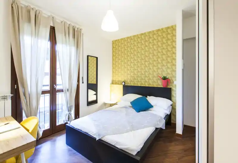 Dormitorio luminoso y bien decorado con cama doble, cojines decorativos y pared de acento con un tono cálido. Gran ventana con cortinas translúcidas que aporta mucha luz natural; escritorio compacto y silla para mayor funcionalidad.
