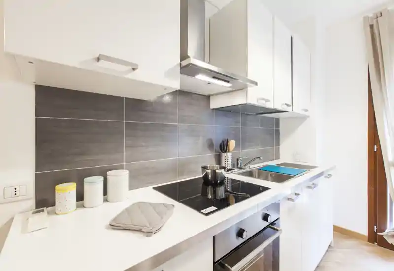 Cuisine compacte moderne avec meubles blancs, plaque à induction, four encastré et évier en acier. Surfaces lumineuses et propres avec de petits objets décoratifs, parfaite pour une annonce locative.