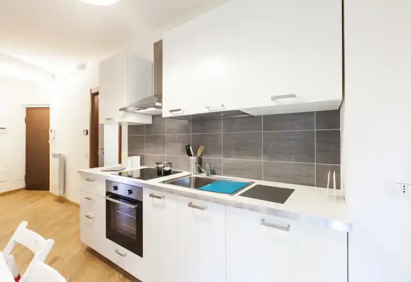 Cuisine moderne et lumineuse avec placards blancs, four encastré et plaque à induction, évier inox et crédence grise — propre et bien éclairée pour un petit appartement.