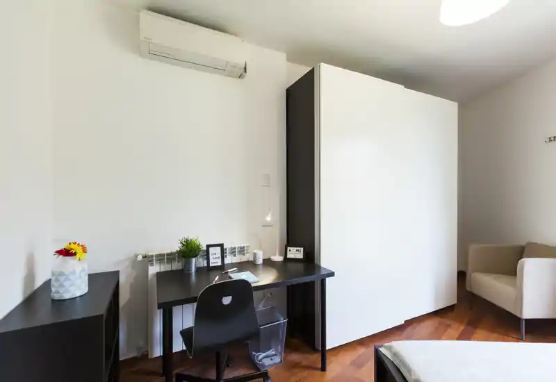 Dormitorio compacto con zona de trabajo: cama individual (vista parcial), escritorio negro con silla y lámpara, armario y butaca; estilo moderno y suelo de madera cálida.