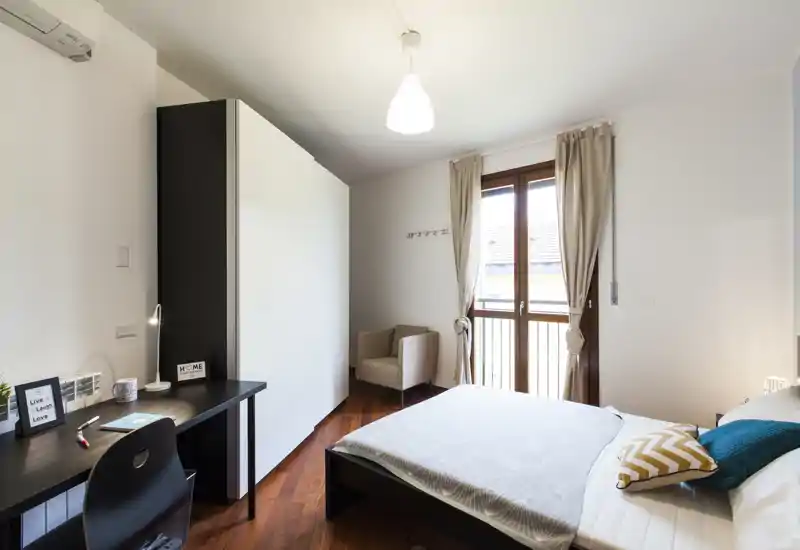 Habitación luminosa y bien presentada con cama doble, cojines decorativos y un escritorio; la luz natural de las puertas al balcón y el suelo de madera aportan calidez.