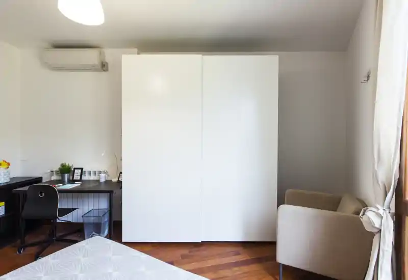 Rincón de dormitorio ordenado con el borde de la cama visible, gran armario blanco, escritorio y un cómodo sillón junto a la ventana — luminoso y práctico para estancias cortas o largas.