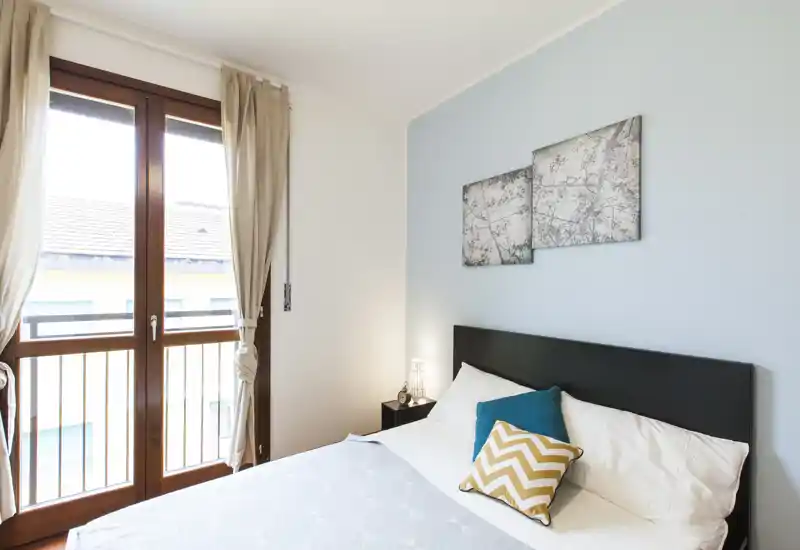 Dormitorio luminoso y moderno con amplia ventana, cojines decorativos y cabecero oscuro — ideal para anuncios que destaquen la luz natural y la comodidad.