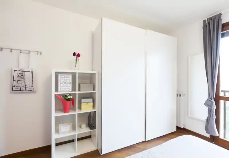 Coin de chambre lumineux et moderne avec une grande armoire blanche, une étagère décorée et un lit partiellement visible. La lumière naturelle venant de la fenêtre crée une atmosphère propre et aérée, parfaite pour les annonces.