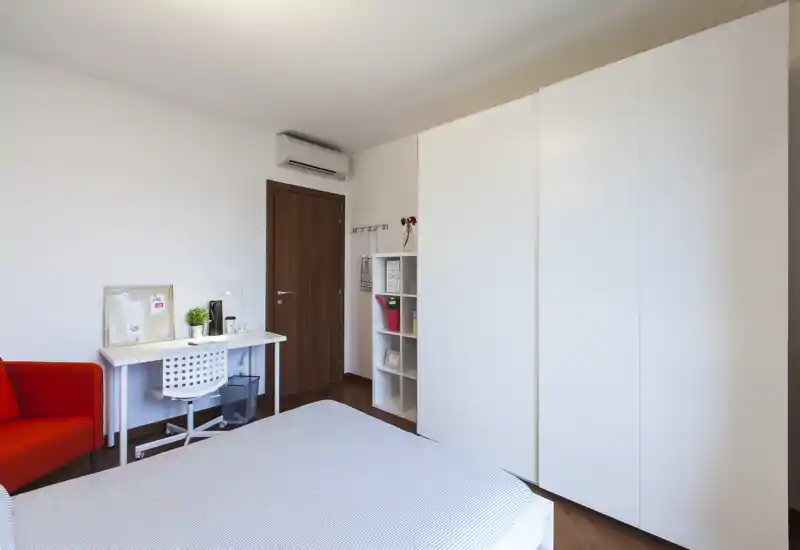 Chambre lumineuse et moderne avec un grand lit au premier plan, une armoire blanche, un petit bureau et un fauteuil rouge — style minimaliste et lumière naturelle abondante.