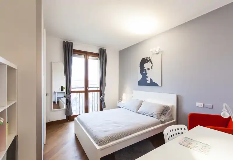 Chambre lumineuse et bien mise en scène avec un lit double centré sur un mur gris, lumière naturelle venant de la porte-fenêtre, lampes de chevet et décoration contemporaine épurée.