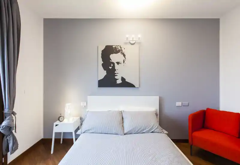 Chambre moderne et lumineuse avec lit double centré, linge de lit neutre, table de chevet et un fauteuil rouge vif qui apporte une touche de couleur.