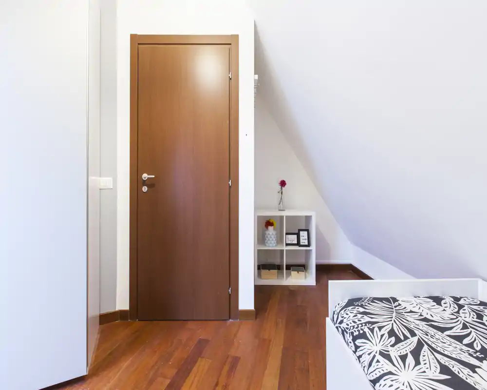 Angolo di camera da letto luminoso e minimale con un letto singolo in primo piano, pavimento in legno caldo e un piccolo scaffale sotto il soffitto inclinato.