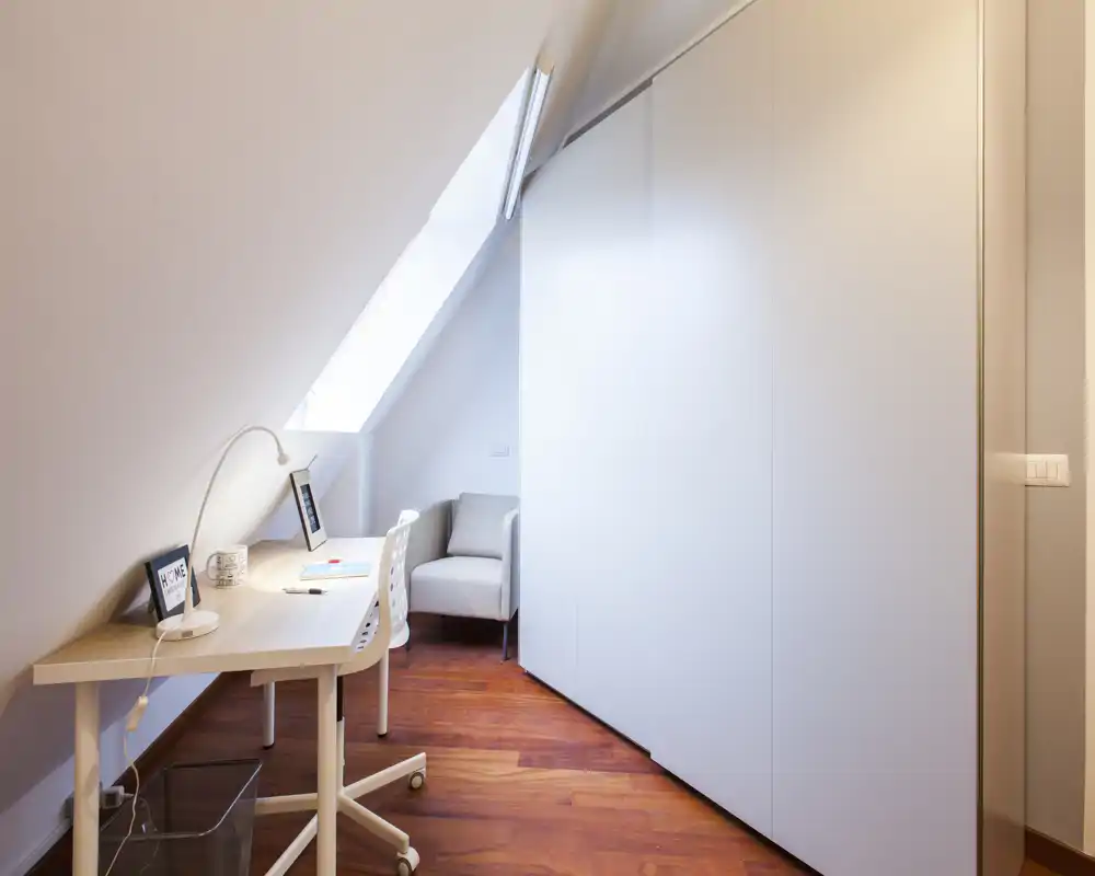 Angolo studio luminoso e minimale in una mansarda, con scrivania bianca, lampada da tavolo e una comoda poltrona accanto a grandi armadi a muro e una finestra a tetto.