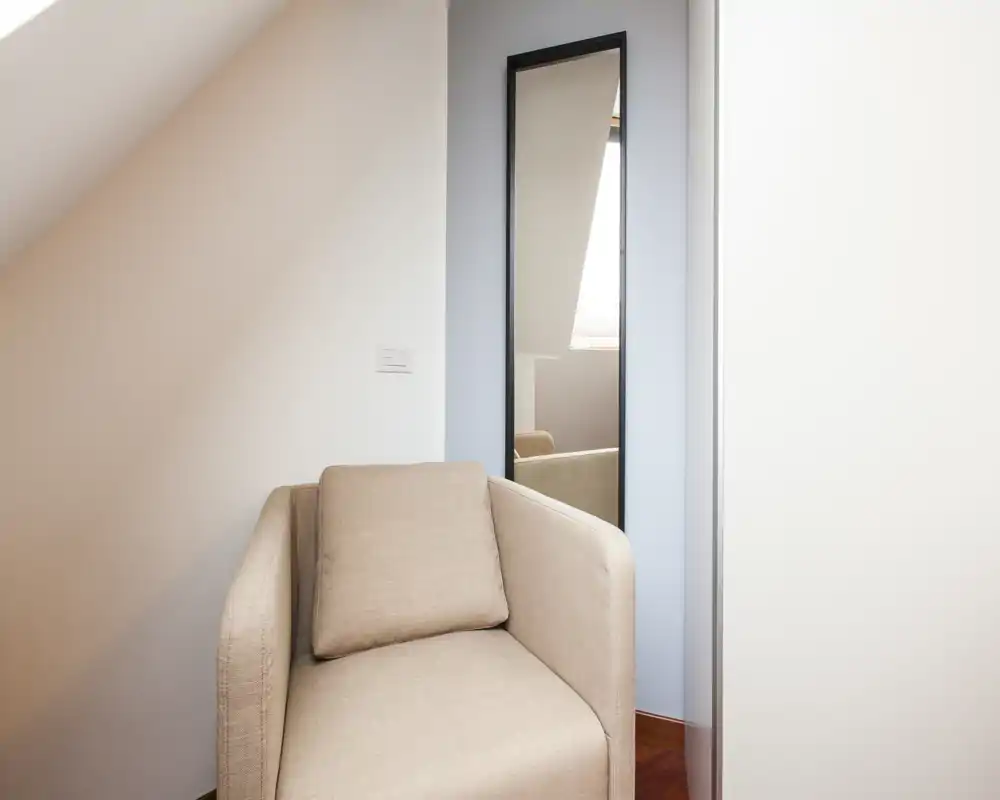 Angolo accogliente con poltrona beige chiaro e specchio a figura intera che riflette luce naturale morbida. Stile minimale e caldo, perfetto come zona lettura o piccolo soggiorno.