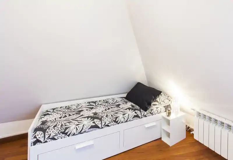 Camera singola con letto contenitore bianco sotto soffitto inclinato, biancheria a motivo e lampada da comodino che creano un ambiente caldo e ordinato.
