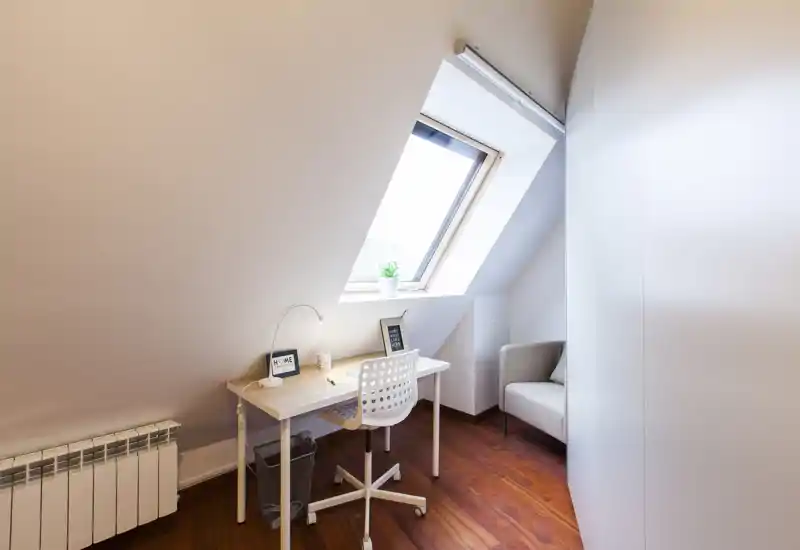 Spazioso angolo studio in mansarda con una scrivania bianca sotto la finestra a tetto, sedia moderna e una poltrona accogliente nell'angolo — perfetto per un home office tranquillo.
