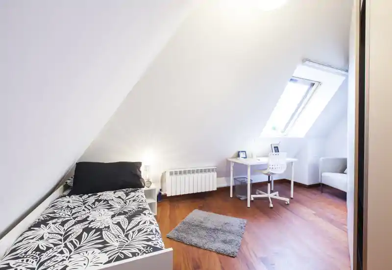 Camera mansardata luminosa con letto singolo, lampada da comodino, scrivania sotto la finestra a tetto e pavimento in legno — uno spazio accogliente e ben illuminato ideale per studenti o ospiti.
