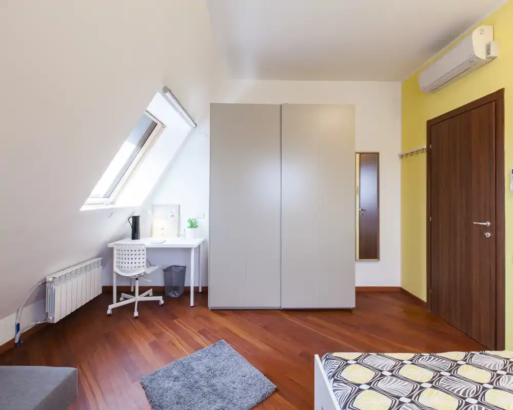 Luminoso camera in mansarda con vista parziale del letto, un grande armadio, una scrivania sotto la finestra a tetto e pavimenti in legno caldo — ariosa e ben illuminata.