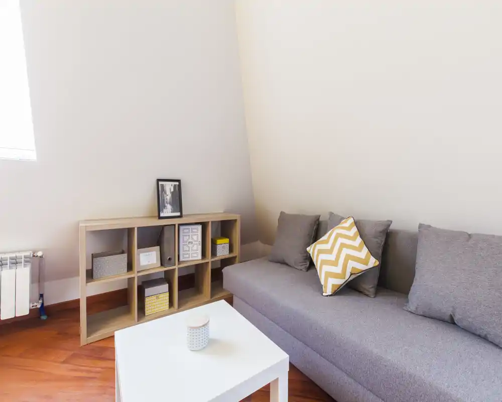 Accogliente soggiorno luminoso con divano grigio, cuscino decorativo, tavolino bianco e scaffalatura a cubi — perfetto per un appartamento compatto.
