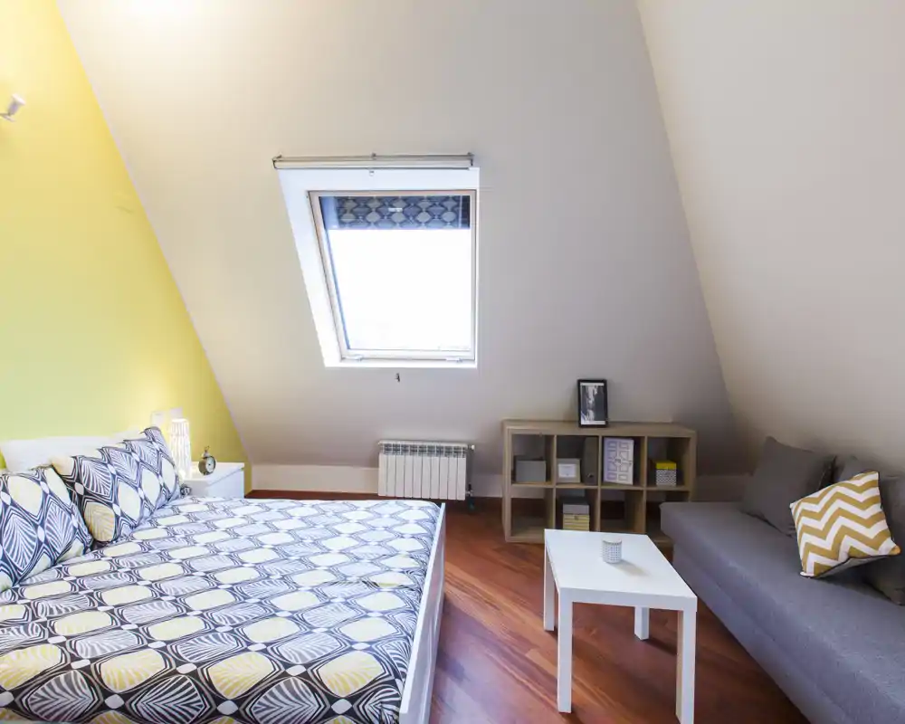 Camera da letto mansardata, luminosa e moderna con copriletto a motivo, finestra a tetto e un piccolo angolo salotto — ideale per soggiorni accoglienti con molta luce naturale.