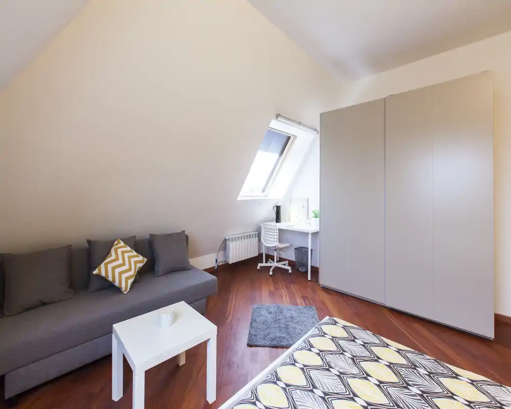 Camera da letto in mansarda luminosa con copriletto a fantasia, divano grigio e scrivania compatta sotto la finestra a tetto — accogliente, moderna e ben allestita per affitti brevi.