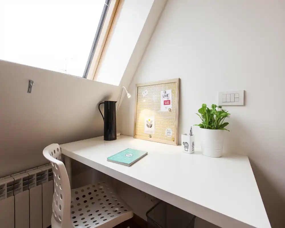Angolo studio in camera da letto sotto una finestra a tetto con scrivania bianca, sedia moderna forata, lampada, bacheca decorativa e pianta: ambiente luminoso, minimale e funzionale.