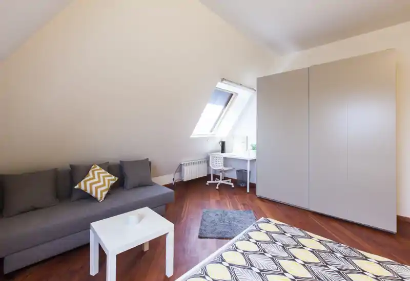 Camera da letto in mansarda luminosa con copriletto a fantasia, divano grigio e scrivania compatta sotto la finestra a tetto — accogliente, moderna e ben allestita per affitti brevi.