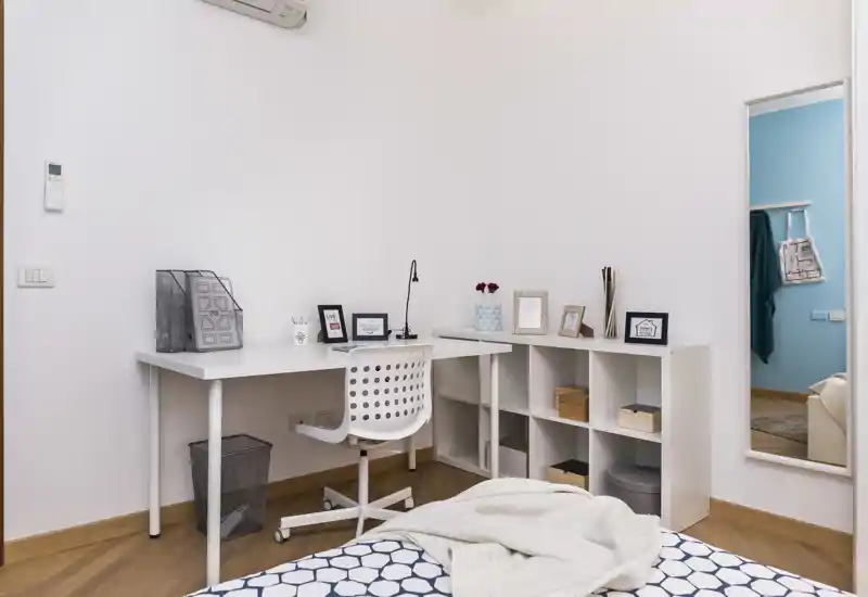 Bright, neatly staged bedroom with a visible bed in the foreground, a white desk and shelving ideal for a work corner, and simple decorative touches that create a calm, modern atmosphere.